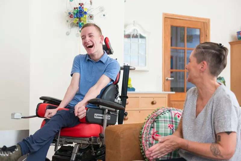 A young man in a powered wheelchair laughing with his caregiver in his living room
